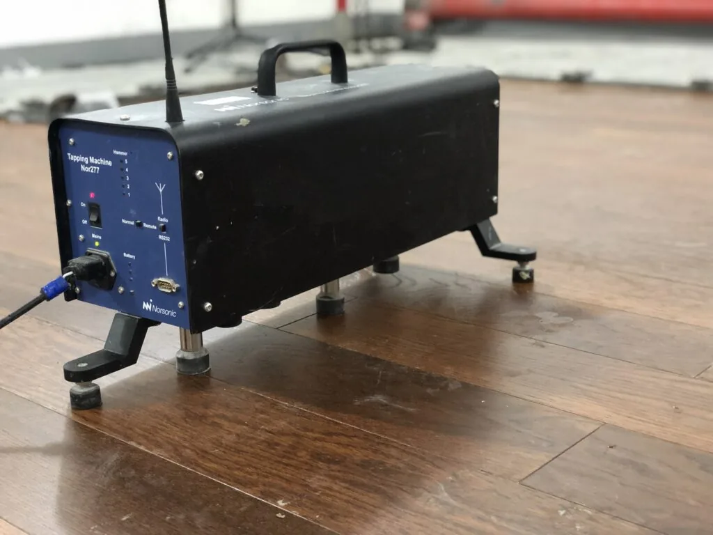 Máquina de impacto normalizada de acústica, con sus martillos calibrados, colocada sobre un piso para medir la transmisión de sonido de impacto hacia el espacio inferior.